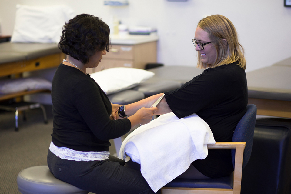 Two women sitting down in a clinic. The physical therapist on the left is treating the elbow of the one on the right.