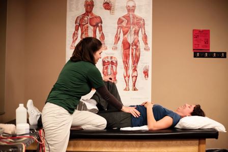 Female physical therapist working on the hips and legs of a female patient as she lies on a table at a clinic underneath a poster illustrating muscular anatomy.