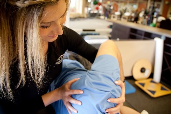 Blonde female physical therapist adjusts a patient's shoulder as they lie on a clinical table.