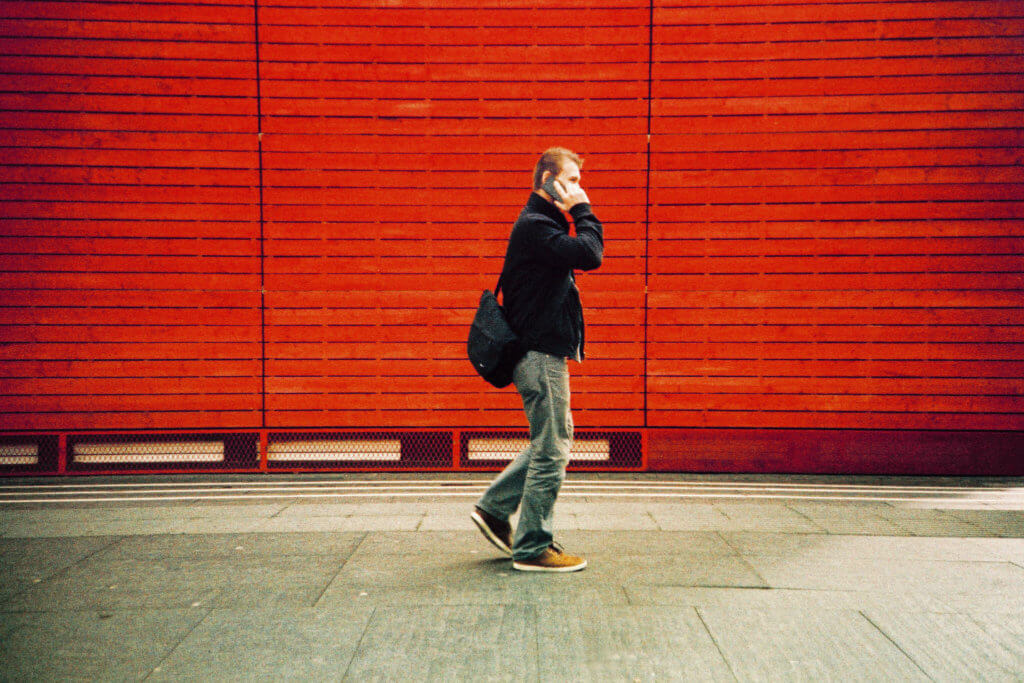 Man walking on a sidewalk while talking on a phone. The wall behind him is bright red.
