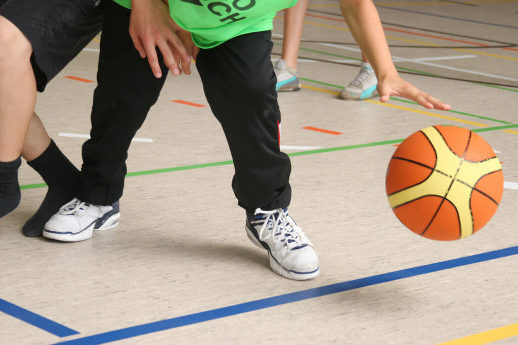 Feet and lower legs of two kids playing basketball at a gym.