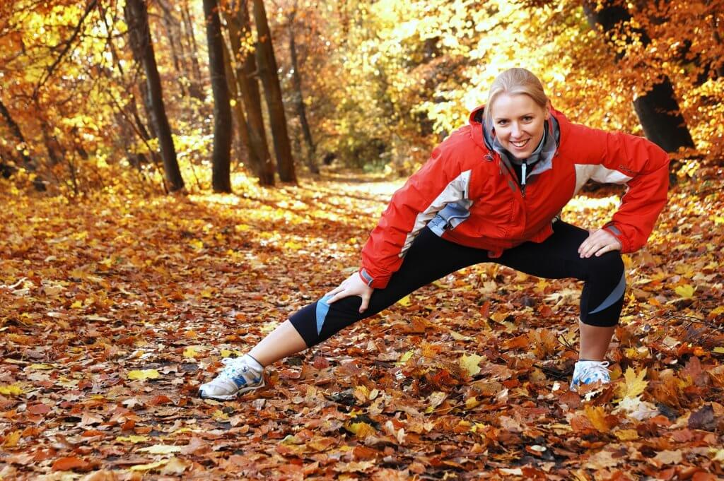 White woman stretching her legs while in a park or forest in the fall, surrounded by trees and yellow and brown leaves on the ground.