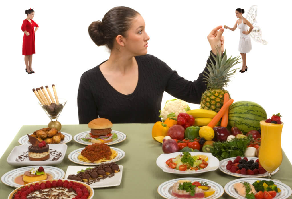 Woman sitting at a dining room table with examples of healthy and not-so-healthy foods. An angel is trying to get her to eat the healthy food, and a devil is trying to get her to eat the unhealthy foods.