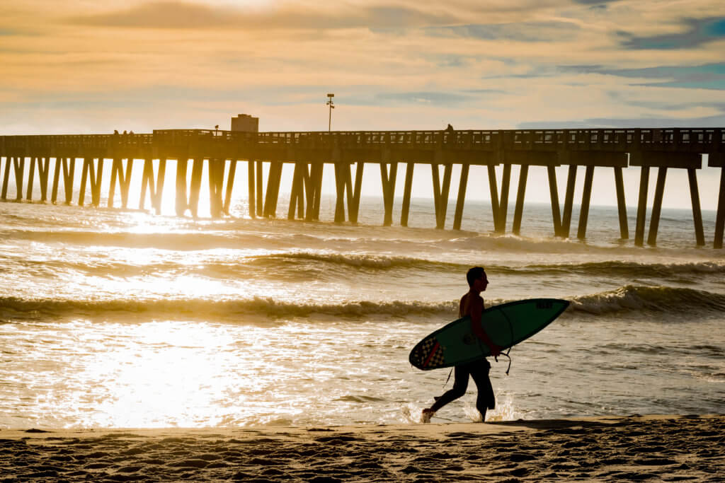 Male surfer carrying his surfboard under his arm as he walks on the beach in the shadows of the pier.