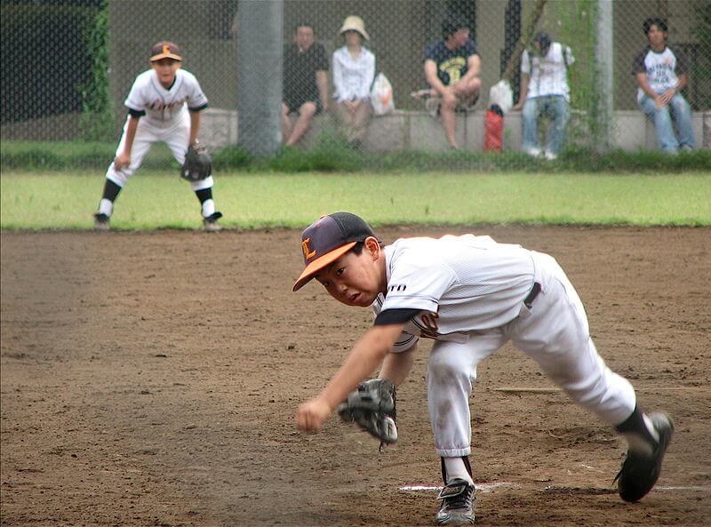 Young baseball pitcher of Asian descent leans toward the ground after releasing a ball toward the batter.