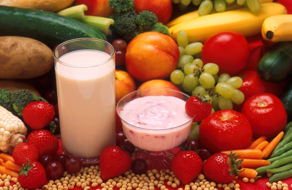 Photograph of glass of milk and bowl of yogurt surrounded by fresh fruits, vegetables and grains.
