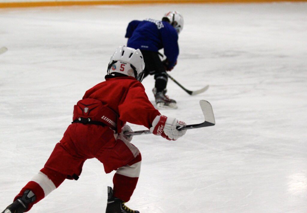 In an ice skating rink, a young hockey player in red skates toward another young hockey player in blue.