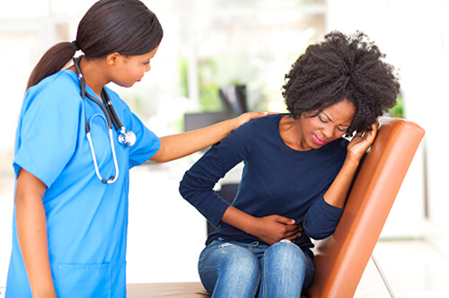 Black female middle aged patient crumples in pain on a seat in a clinic. A Black healthcare provider touches her on her shoulder and expresses concern.