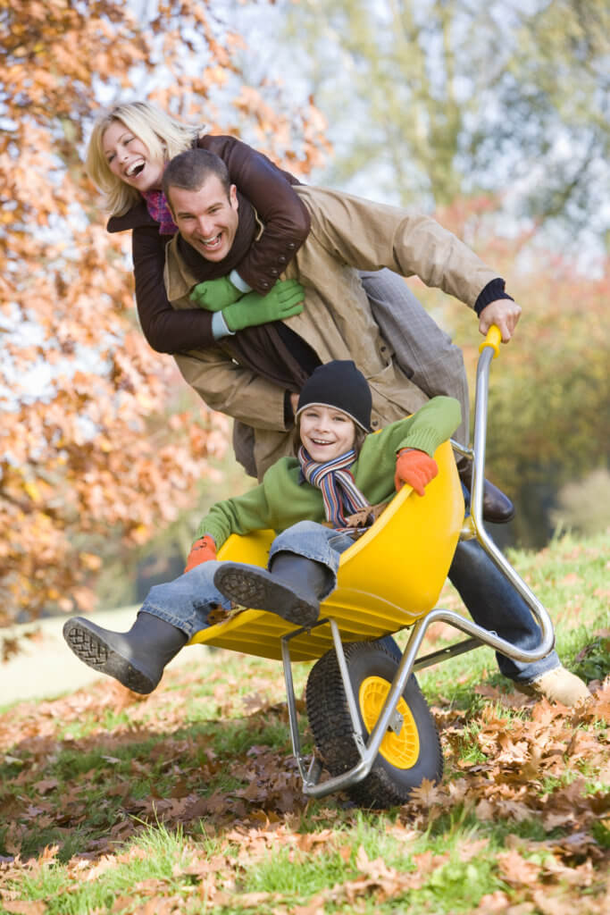 A family of a mom, dad and child play with a wheelbarrow in the yard.