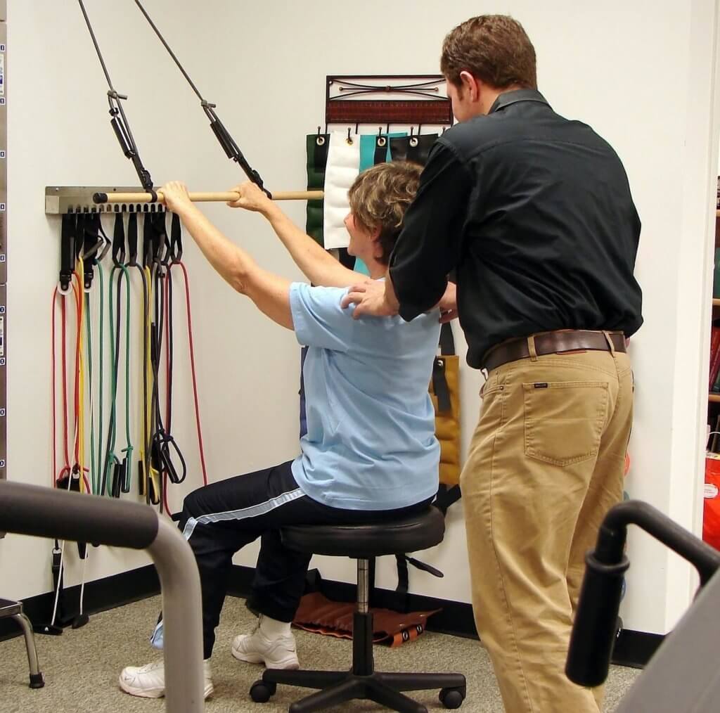 A male physical therapist pushes down on a femal patient's shoulders as she pulls on a bar attached to elastic resistance bands.