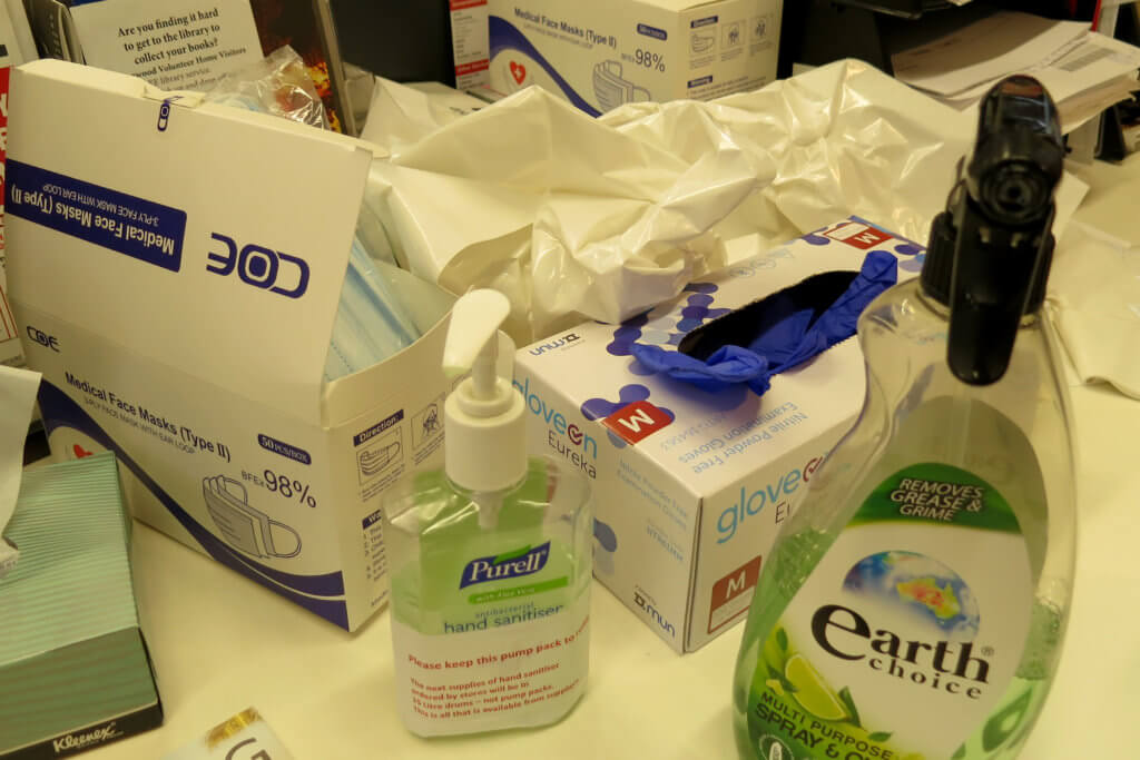 Photograph of several cleaning supplies for COVID on a table: Sanitizer, plastic gloves, face masks, cleaning spray, and more.