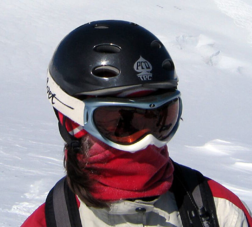 Photograph of the head of a person with heavy snow gear while standing on a snowy mountain: Ski helmet and goggles, neck gaiter, and heavy jacket.