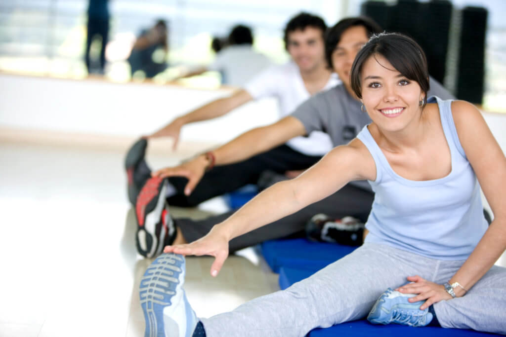 Woman and two men stretching their legs while sitting on a mat at the gym.