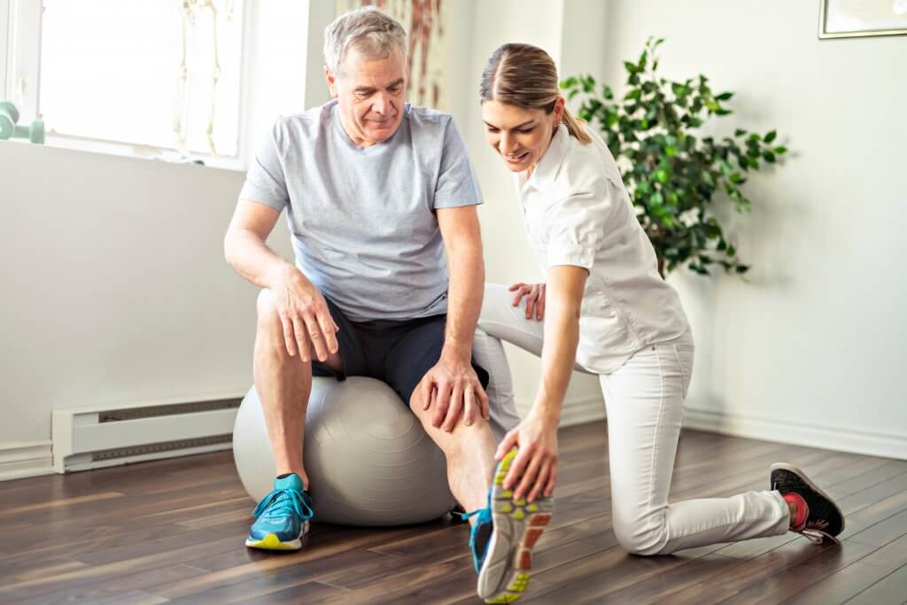 Older male patient working with a younger female physical therapist to stretch his left calf and hamstring while sitting on a large exercise ball.